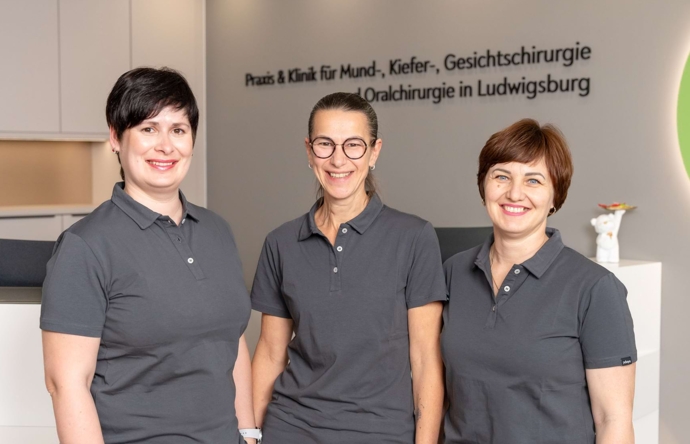 Bild dreier Mitarbeiterinnen, die vor dem Empfangsthresen stehen und in die Kamera lächeln