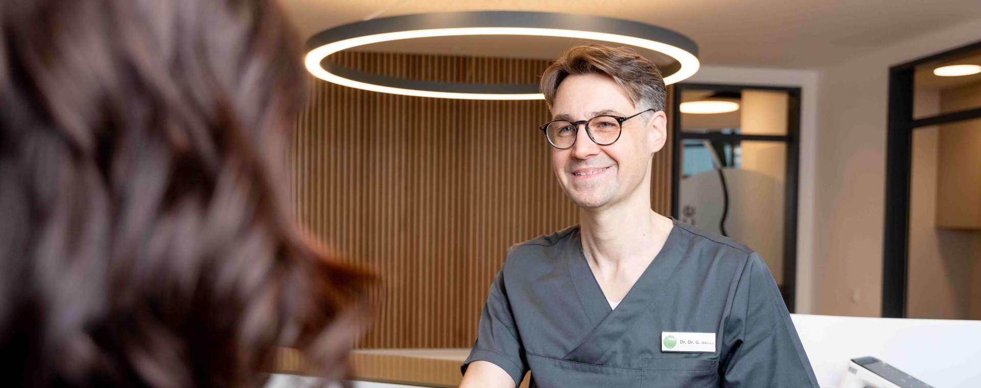 Dr. Dr. Santo streckt seinen Arm im Foyer der Praxisklinik aus, um Patient:innen zu begrüßen